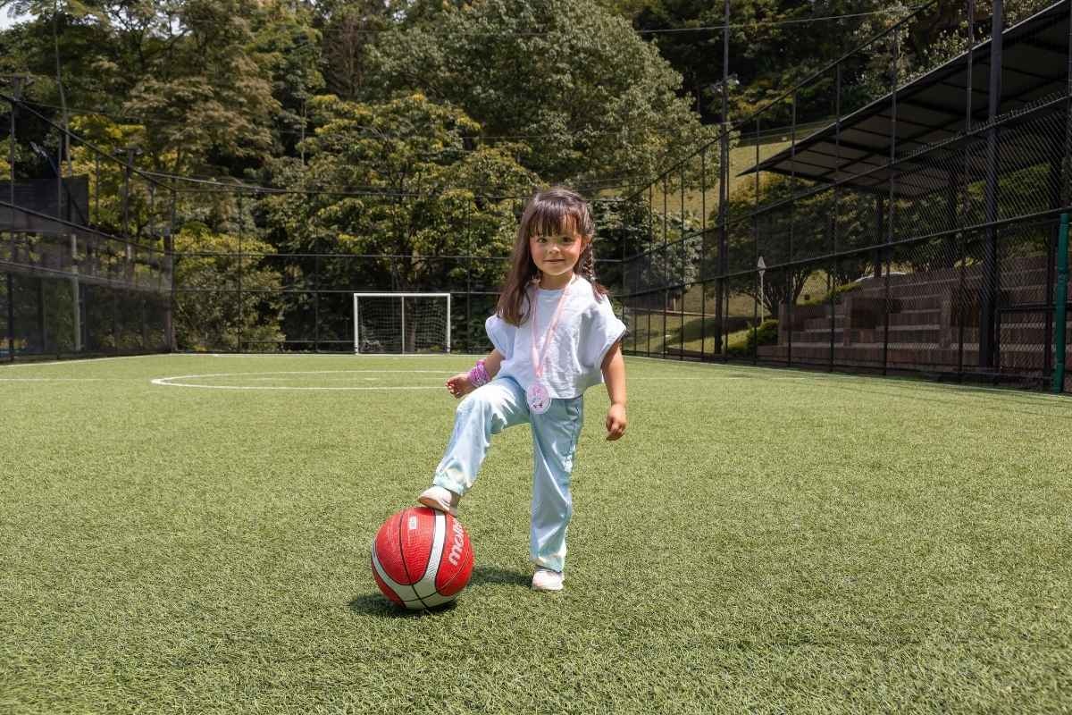 Niña pequeña con conjunto de ropa deportiva celeste posando con un balón en una cancha de fútbol de césped sintético.