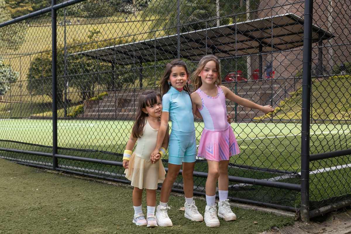 Tres niñas sonriendo en una cancha deportiva usando ropa deportiva infantil en colores pastel amarillo, celeste y rosa