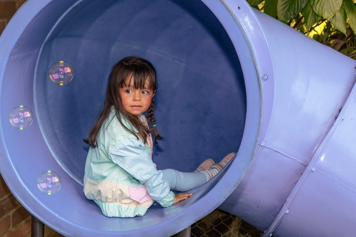 Niña pequeña con chaqueta ligera y leggings cómodos sentada en un túnel de juegos de jardín infantil.