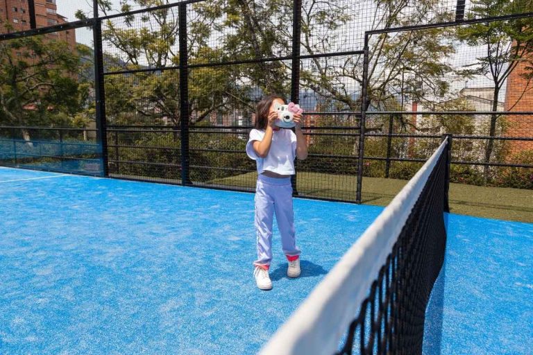 Niña pequeña con pantalón deportivo lila y camiseta blanca usando una cámara de juguete en una cancha de pádel