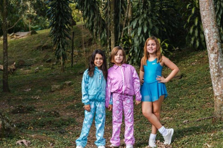Tres niñas posando en un entorno natural con las últimas tendencias en ropa infantil: conjuntos deportivos en azul y rosa con acabados satinados y un vestido deportivo celeste.